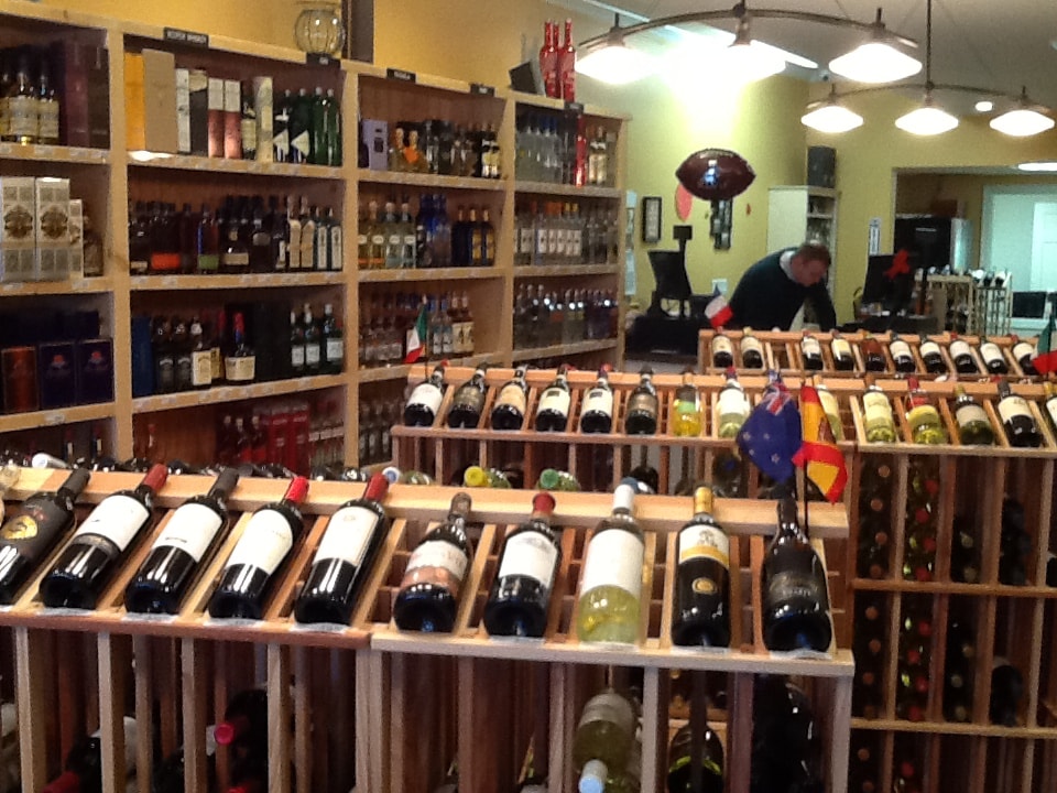 The image shows a well-stocked wine shop with numerous bottles displayed on shelves, along with signage indicating different types of wine.