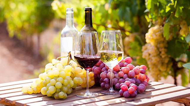 Bottle of wine and glasses on table with bunches of grapes and wine bottles behind them.