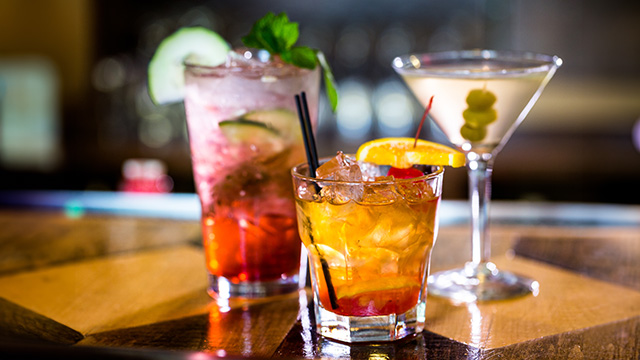 The image shows three cocktails on a bar counter with garnishes, accompanied by a slice of orange.